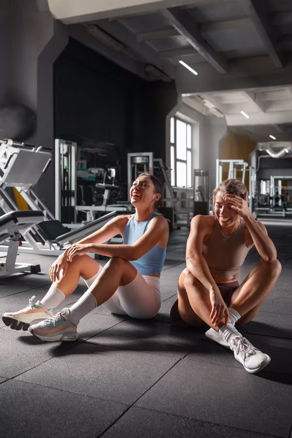 Two women in designer athleisure sharing a laugh on the gym floor, capturing the social aspect of minimalist wellness.